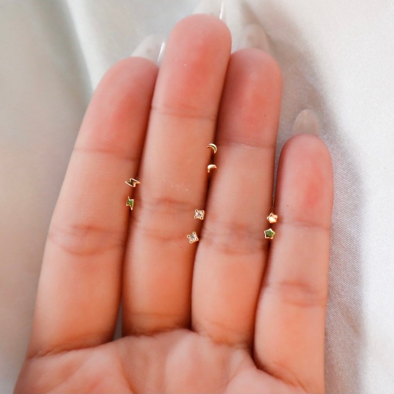 May include: A set of five gold earrings with different designs, including a crescent moon, a lightning bolt, a star, and two square-shaped earrings with small clear stones.