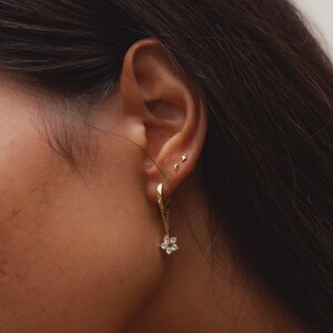 May include: Gold hoop earrings with a dangling flower charm featuring clear cubic zirconia stones. The earrings are shown in a person's ear.