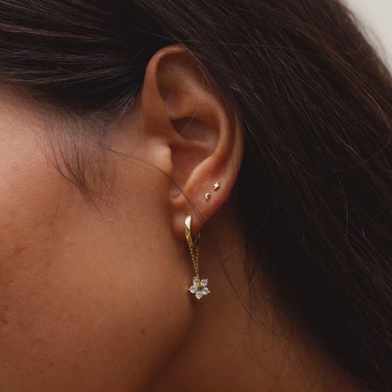 May include: Gold hoop earrings with a dangling flower charm featuring clear cubic zirconia stones. The earrings are shown in a person's ear.