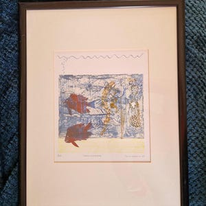 Op de afbeelding: Gelijst drukwerk van een blauw en geel abstract kunstwerk met twee rode en blauwe vissen. De print is getiteld "Hidden Waters" en gesigneerd door de kunstenaar "Jane Wadham 83".