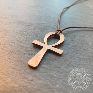 Könnte beinhalten: Eine goldfarbene Ankh-Halskette mit einem braunen Lederband. Der Anhänger ist in Deutschland von Gem Sessions hergestellt.