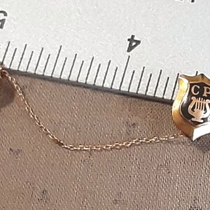 May include: A gold-colored chain connects two pins. One pin features a black shield with the letters "CP" and a lyre inside. The other pin is a gold-colored lantern with the number "29" on it.