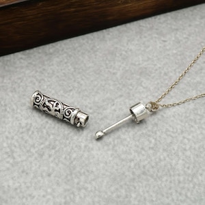 May include: A silver pendant necklace featuring a cylindrical charm with intricate patterns. The charm is detached, showing its internal mechanism. The necklace has a fine chain and a small, rounded tip.