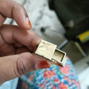 May include: A gold-colored ring with a small, rectangular compartment that slides open. The ring is held in a hand with painted nails, and the background is blurred.