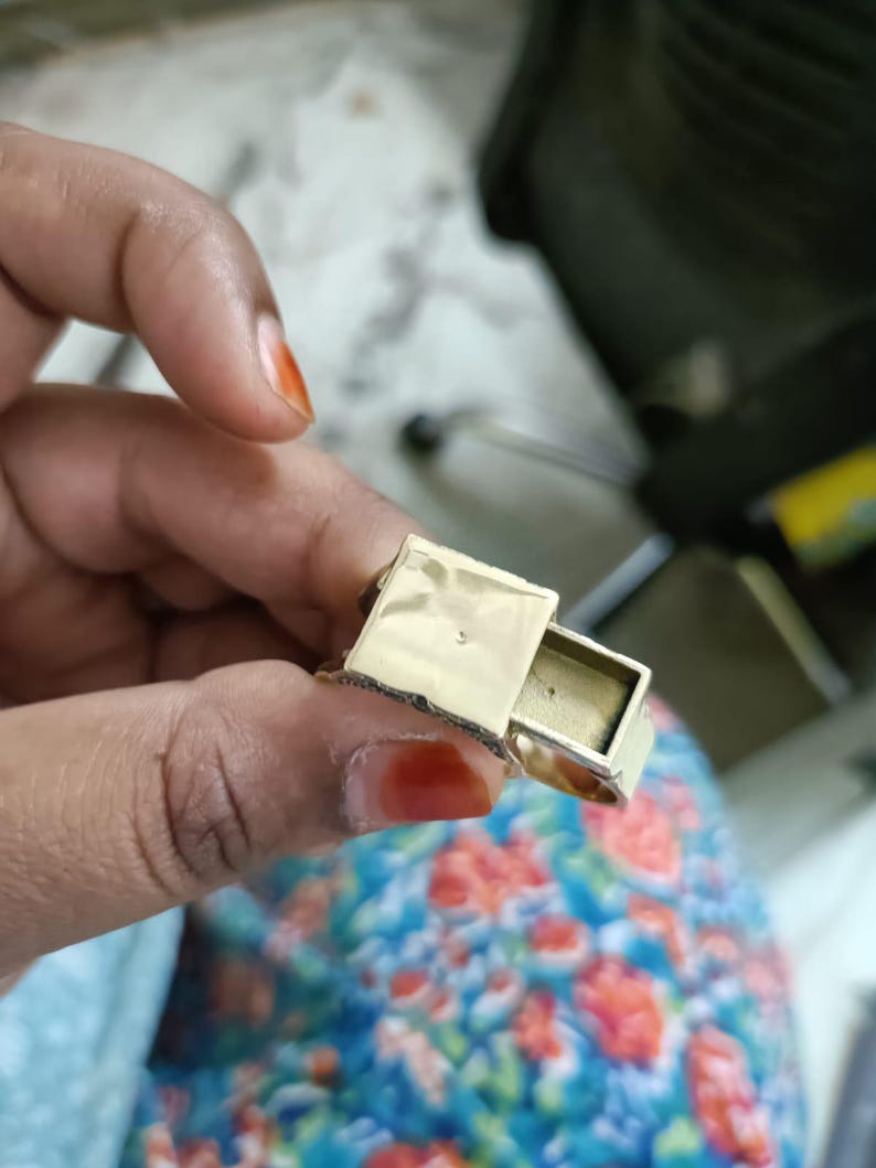 May include: A gold-colored ring with a small, rectangular compartment that slides open. The ring is held in a hand with painted nails, and the background is blurred.