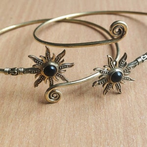 Könnte beinhalten: Zwei goldfarbene Armbänder mit schwarzen Edelstein-Akzenten in Sonnenform. Die Armbänder sind aus dünnem Draht gefertigt und haben ein spiralförmiges Design am Ende.