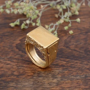 May include: A gold-colored ring with a rectangular top and ornate detailing on the side. The ring has a hinged compartment. The ring is set against a wooden background with a sprig of greenery.