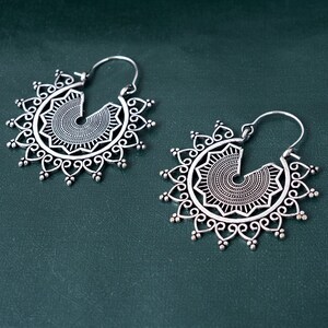 May include: A pair of silver hoop earrings with intricate, circular designs. The earrings have a mandala-like pattern with a central circle and radiating spokes.