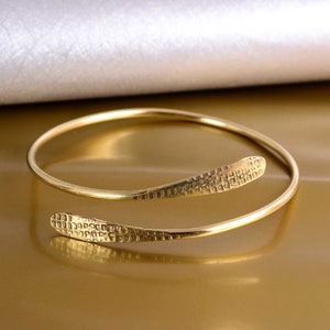 May include: A gold-toned bangle bracelet with a textured, patterned design. The bracelet is open and has a curved shape.