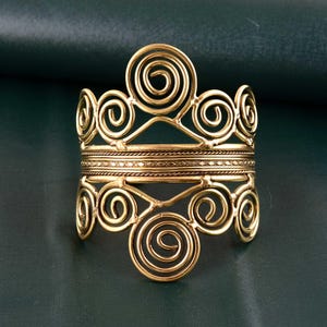 Puede incluir: Un brazalete de puño dorado con un diseño intrincado que presenta múltiples patrones de espirales y remolinos. El brazalete tiene una banda ancha con líneas horizontales detalladas y elementos decorativos. El diseño general es simétrico y ornamentado, con una estética vintage o antigua.