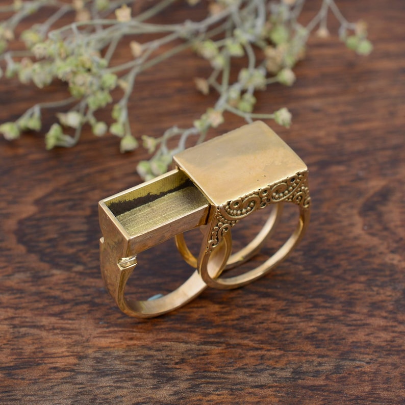May include: A gold-colored ring with a rectangular compartment that slides open, revealing a small interior. The ring has an ornate design on the side and two bands for the fingers. The ring is set against a wooden background.