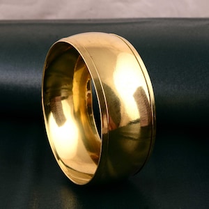 May include: A gold-colored bangle bracelet with a smooth, polished surface. The wide band has a simple, elegant design, reflecting light and creating a warm, lustrous appearance. The jewelry piece is a classic accessory.