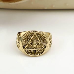 Può includere: Un anello color oro con un design centrale di un occhio all'interno di un triangolo, circondato da raggi di sole. L'anello ha una superficie dettagliata e strutturata, probabilmente in metallo. Il design dell'anello suggerisce un tema mistico o simbolico.