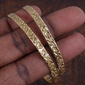 May include: Two gold-tone bangles with a floral design. The bangles are stacked on top of each other.