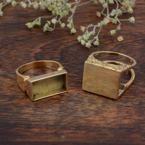 May include: Two gold-colored rectangular ring blanks on a dark wood surface. One ring has a plain rectangular setting, while the other features an ornate design on the setting. Dried flowers are in the background.