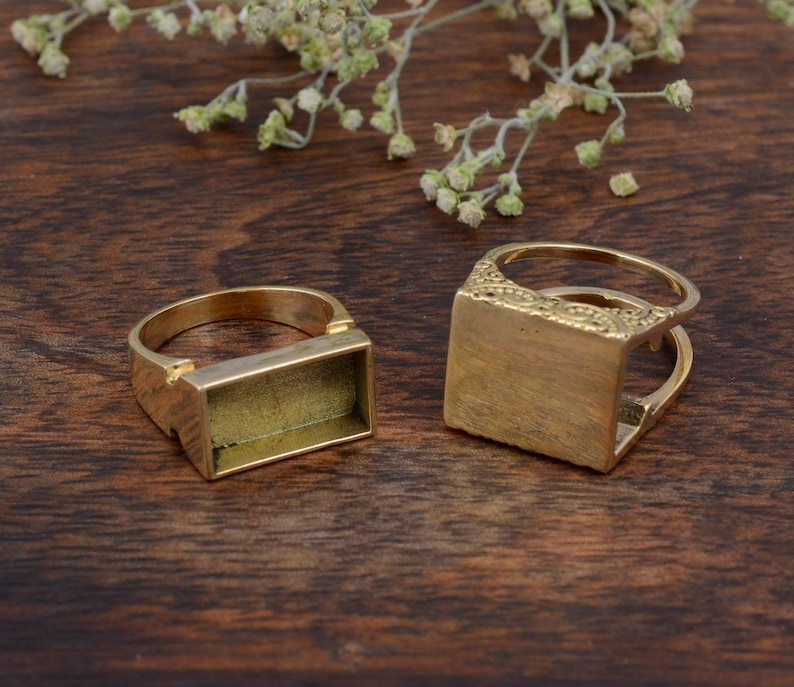 May include: Two gold-colored rectangular ring blanks on a dark wood surface. One ring has a plain rectangular setting, while the other features an ornate design on the setting. Dried flowers are in the background.