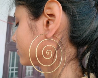 May include: A large, circular gold-toned earring featuring a continuous spiral design. The metal surface has a subtle hammered texture, creating a reflective finish. The earring is shaped as a thin, open wire coil that loops outward from the center.