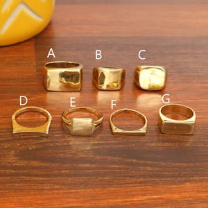 May include: A collection of seven gold-toned rings labeled A through G. The rings feature various band widths and face shapes, including rectangular, square, and rounded designs. Each ring has a smooth, polished finish and a metallic gold appearance.
