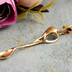 Puede incluir: Un collar con un colgante de oro en forma de cráneo de pájaro. El colgante tiene forma de cuchara con un cráneo hueco en la parte superior. El collar está en una cadena de oro delicada.
