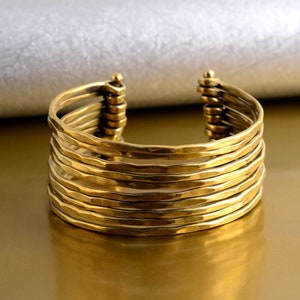 Könnte beinhalten: Ein goldfarbenes Metall-Armreif mit gehämmerter Oberfläche. Das Armband besteht aus mehreren übereinander gestapelten Bändern.