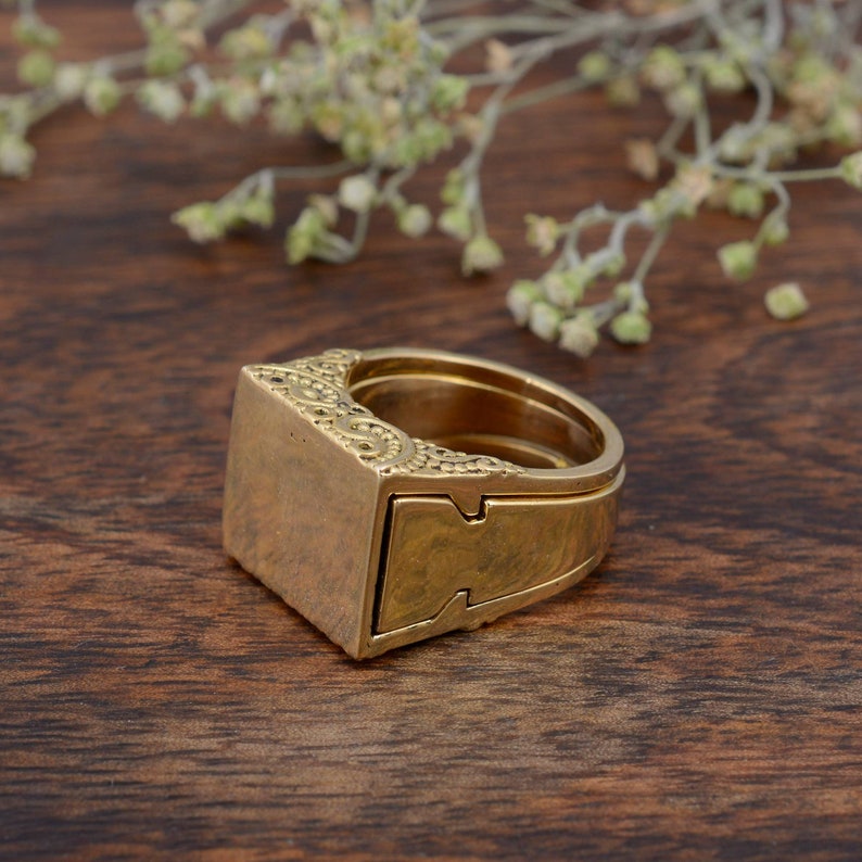 May include: A gold-colored ring with a rectangular top and intricate decorative patterns. The ring has a unique design with a sliding compartment. The ring is set against a dark wooden surface.