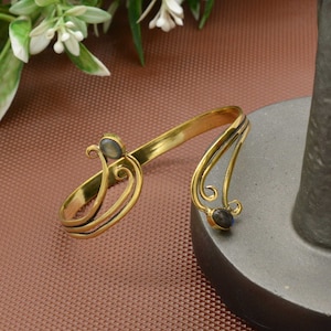 Peut inclure: Un bracelet de bras doré avec un design ouvert. Le bracelet présente une forme incurvée avec des volutes décoratives et deux petites pierres de forme ovale. Le métal a un aspect légèrement vieilli, et le design est complexe et détaillé.