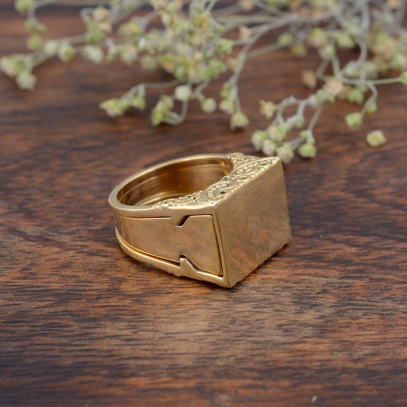 May include: A gold-colored ring with a rectangular top and intricate detailing. The ring has a unique design with a hidden compartment. The ring is set against a dark wooden surface with a blurred floral element in the background.