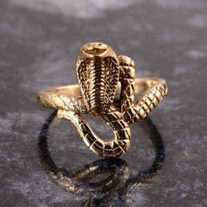 May include: A gold-toned ring featuring a detailed cobra snake design. The snake is coiled with its head raised and its scales are visible.