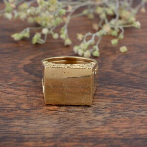 May include: A gold-colored signet ring with a rectangular face and ornate detailing. The ring is set against a dark wood surface, with a blurred floral element in the background. The ring's design is classic and elegant.