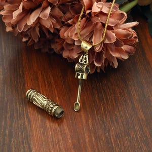 Op de afbeelding: Een goudkleurige metalen hanger aan een ketting met een afneembare cilindrische dop met ingewikkelde reliëfpatronen. De hanger bevat een klein, slank lepelvormig hulpmiddel aan het oogje, hangend aan een bijpassende goudkleurige ketting.