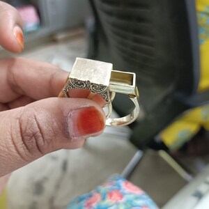 May include: A gold-colored ring with a rectangular compartment that slides open. The ring has an ornate design on the sides of the rectangular compartment. The ring is held up by a person's hand.