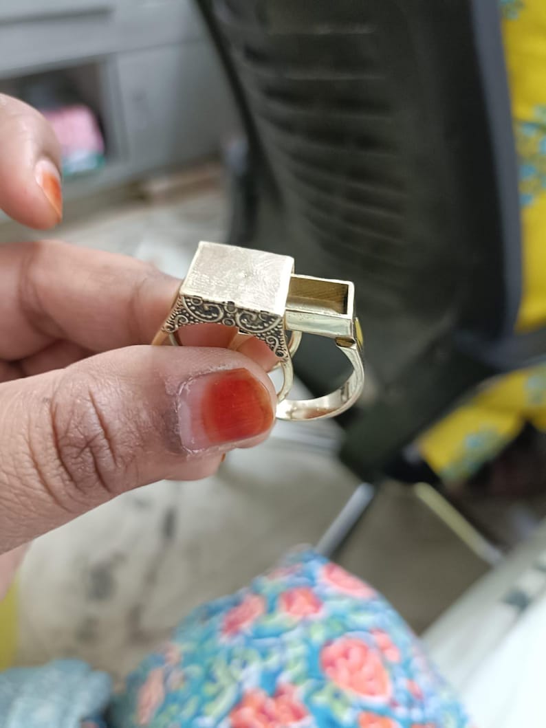 May include: A gold-colored ring with a rectangular compartment that slides open. The ring has an ornate design on the sides of the rectangular compartment. The ring is held up by a person's hand.