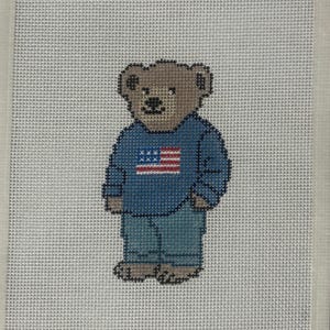 Op de afbeelding: Kruissteekontwerp van een teddybeer met een blauwe trui met een Amerikaanse vlag en een spijkerbroek. De beer is bruin met zwarte details, op een witte achtergrond.