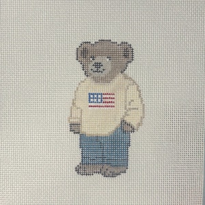 May include: Needlepoint design of a teddy bear wearing a cream-colored sweater with an American flag and blue jeans. The bear is standing on a light beige background. The design is made with cross-stitch.