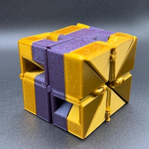 Puede incluir: Un cubo de rompecabezas geométrico dorado y morado con una superficie texturizada. El cubo está hecho de varias piezas que se pueden girar y reorganizar.