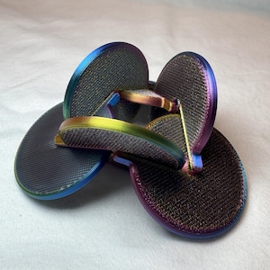 Puede incluir: Un conjunto de chanclas iridiscentes con una superficie texturizada. Las chanclas tienen un color base oscuro con bordes que cambian entre azul, morado y dorado. Las correas también son iridiscentes, creando un efecto colorido.