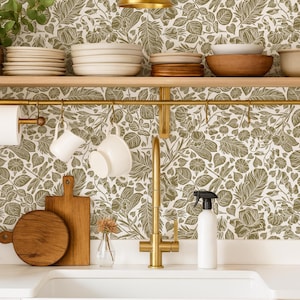 May include: A kitchen scene with a gold faucet, white sink, and floral wallpaper. Wooden shelves hold stacks of plates and bowls. White mugs hang from a gold bar, and a cutting board rests nearby. A white spray bottle sits on the counter.
