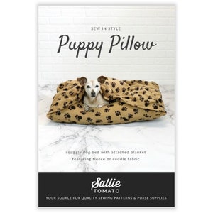 May include: A tan and brown dog bed with a paw print pattern, featuring a small dog nestled inside. The text "Puppy Pillow" is displayed above the bed, with additional text indicating it's a snuggly bed with a fleece blanket.
