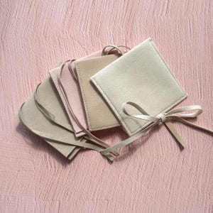 Puede incluir: Cuatro bolsas de fieltro beige y rosa con un lazo. Las bolsas están apiladas una encima de la otra.