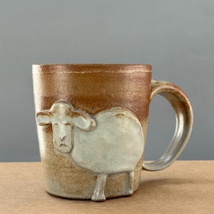 May include: A handmade ceramic mug with a sheep design. The mug is a warm brown color with a lighter beige sheep relief. The handle is a curved, semi-circular shape. The mug is suitable for hot or cold beverages.