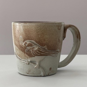 Puede incluir: Taza de cerámica hecha a mano con diseño de pájaro. La taza presenta una combinación de colores marrón claro y blanco, con un relieve detallado de un pájaro posado en una rama. El asa es curvada y tiene un aspecto rústico.