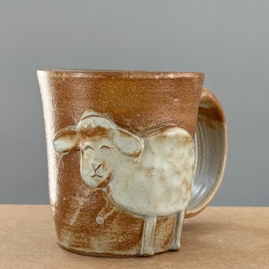 May include: A brown ceramic mug with a sculpted sheep design. The sheep is light beige and has a smiling face. The mug has a large handle and a textured surface.