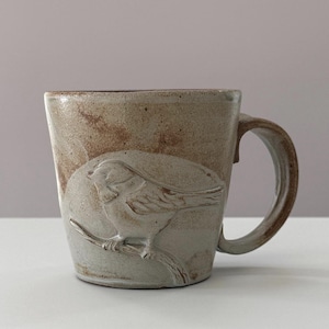 Puede incluir: Una taza de cerámica hecha a mano con un diseño en relieve de un pájaro. La taza presenta una combinación de colores marrón claro y beige con un pájaro detallado posado en una rama. El asa es robusta y complementa la estética rústica. Una pieza única para café o té.