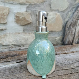 Può includere: Un dispenser di sapone in ceramica con base bulbosa caratterizzato da uno smalto verde salvia lucido e screziato che cola su un corpo in argilla color bianco sporco con macchioline. Il dispenser è dotato di una pompa in acciaio inossidabile.