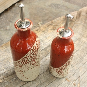 May include: Two ceramic oil and vinegar bottles with red and white speckled glaze and silver pour spouts. The bottles are on a wooden surface.