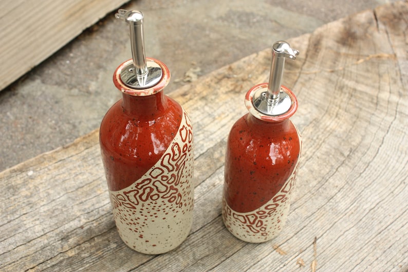 May include: Two ceramic oil and vinegar bottles with red and white speckled glaze and silver pour spouts. The bottles are on a wooden surface.