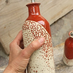 May include: A ceramic olive oil bottle with a red and white speckled glaze. The bottle has a unique design with a red and white pattern. The bottle has a metal spout.