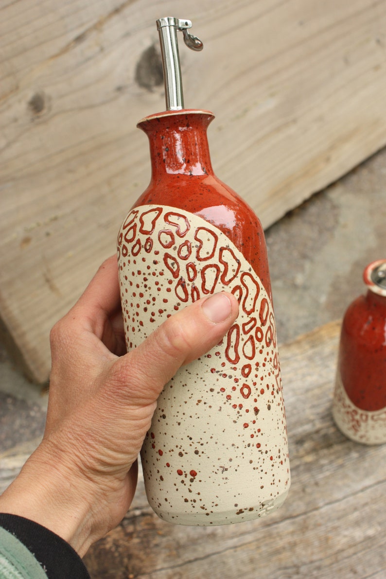 May include: A ceramic olive oil bottle with a red and white speckled glaze. The bottle has a unique design with a red and white pattern. The bottle has a metal spout.
