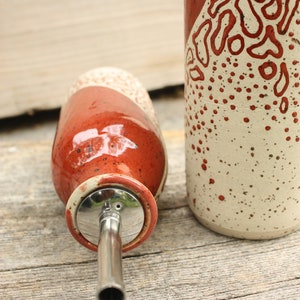May include: Two ceramic oil and vinegar bottles with a red and white speckled glaze. The bottle on the left has a stainless steel pour spout.