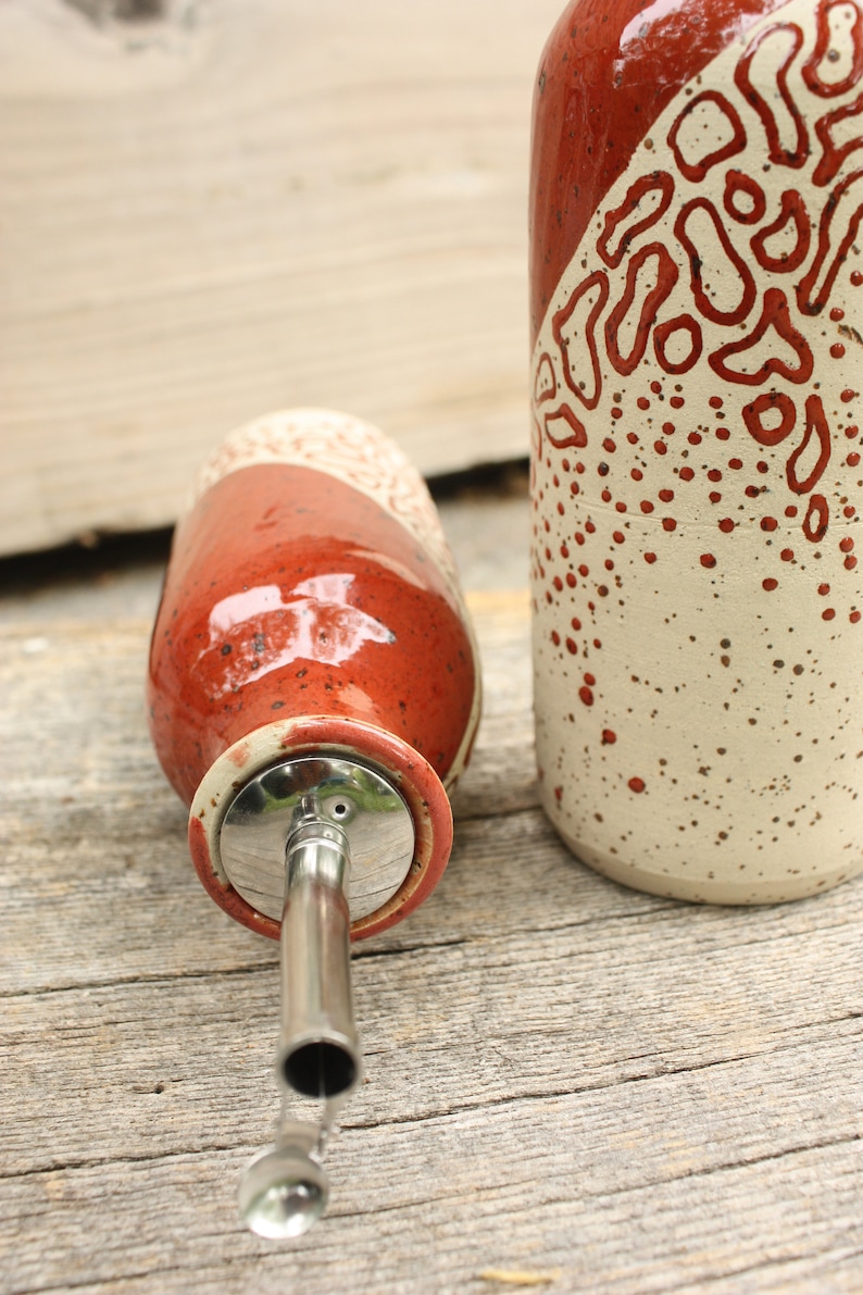 May include: Two ceramic oil and vinegar bottles with a red and white speckled glaze. The bottle on the left has a stainless steel pour spout.